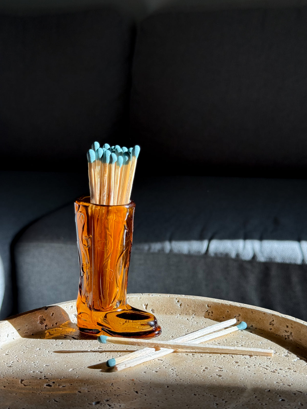 An amber glass match holder filled with wooden matches topped with blue tips, placed on a speckled beige tray in front of a dark gray sofa.
