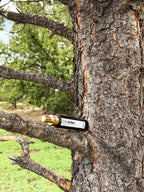 A small amber glass bottle with a gold cap and a white label reading 'Membribar' rests on a tree branch outdoors against a tree trunk.