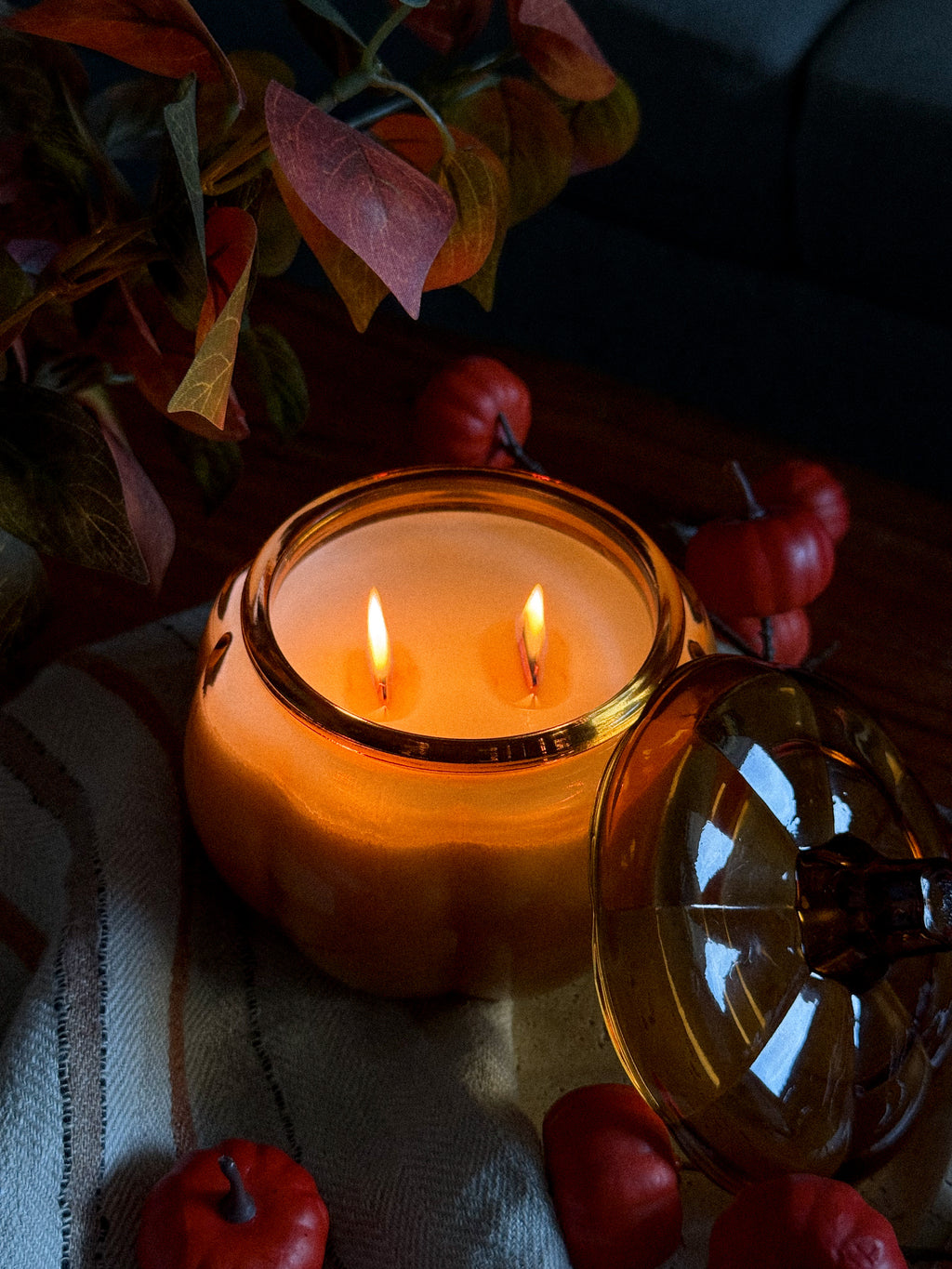 A lit amber glass jar candle with two wicks sits on a striped fabric surface, surrounded by small red decorative pumpkins and autumn leaves in a dimly lit setting.