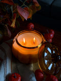 A lit amber glass jar candle with two wicks sits on a striped fabric surface, surrounded by small red decorative pumpkins and autumn leaves in a dimly lit setting.