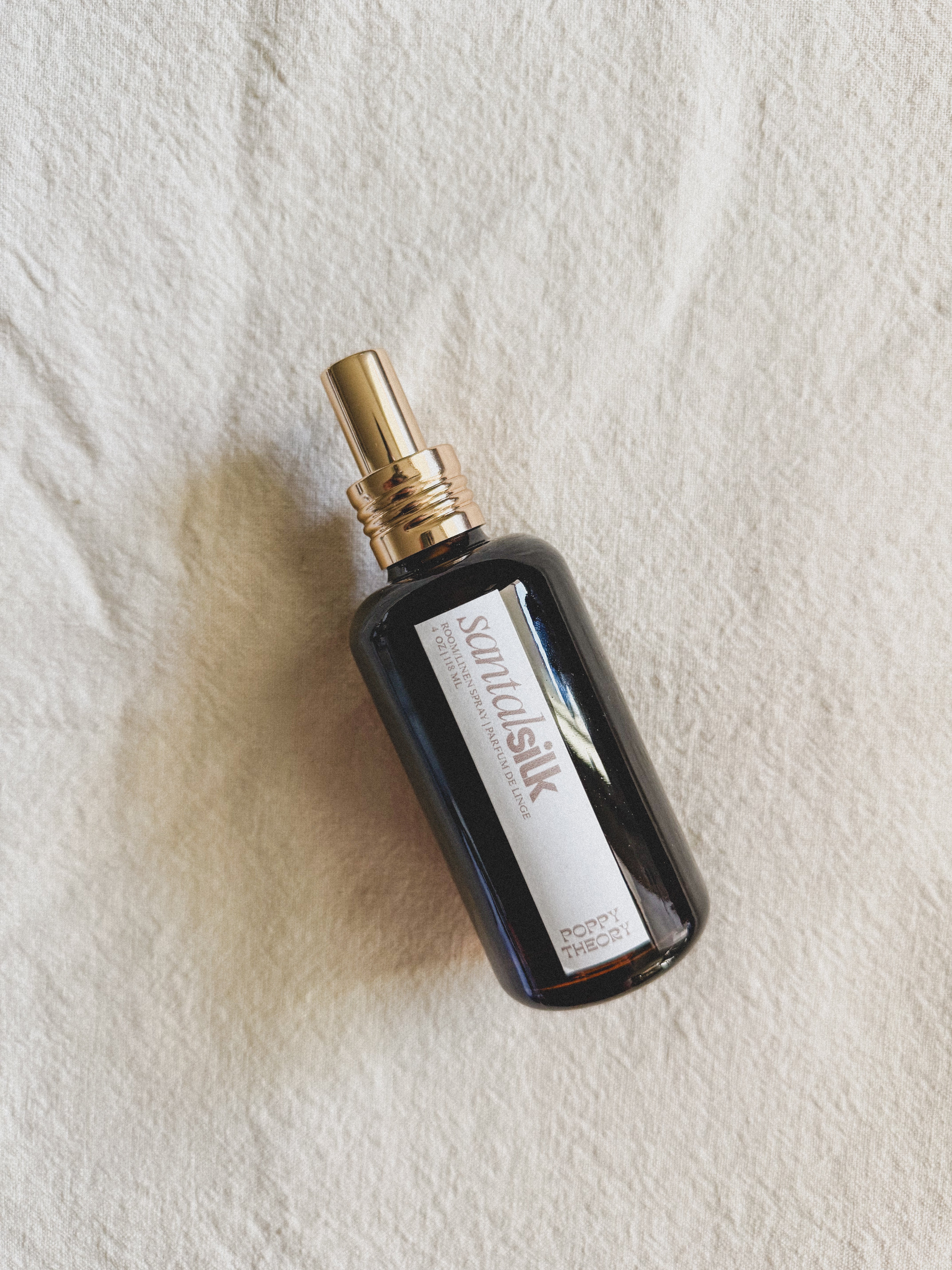 A bottle of Santal Silk Room Spray on a textured off-white fabric background. The bottle is dark amber glass with a metallic gold spray nozzle and a white label featuring the Poppy Theory brand name.