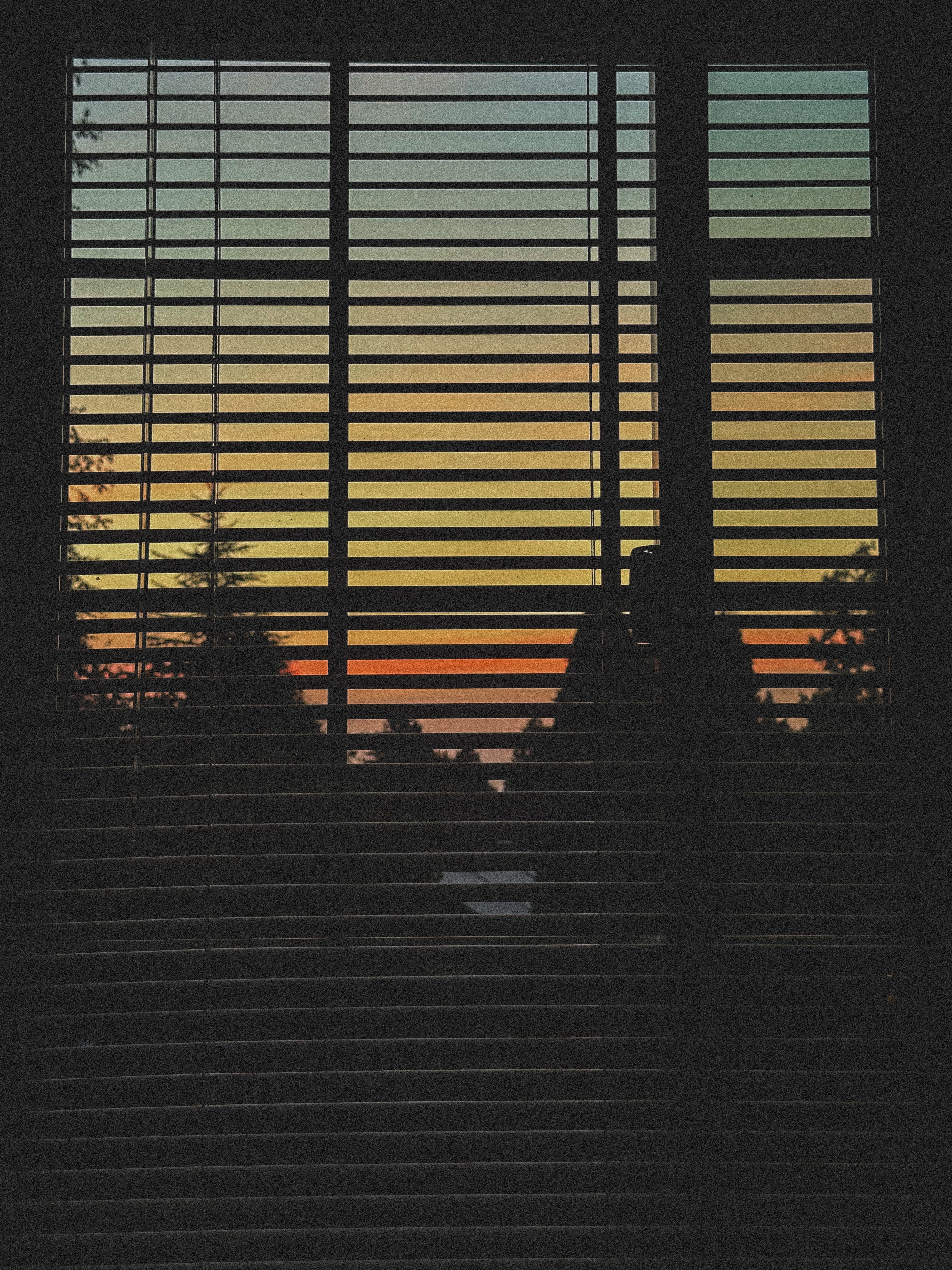 A square art print depicting a sunset seen through horizontal window blinds, with a silhouetted figure standing in front of the glass.